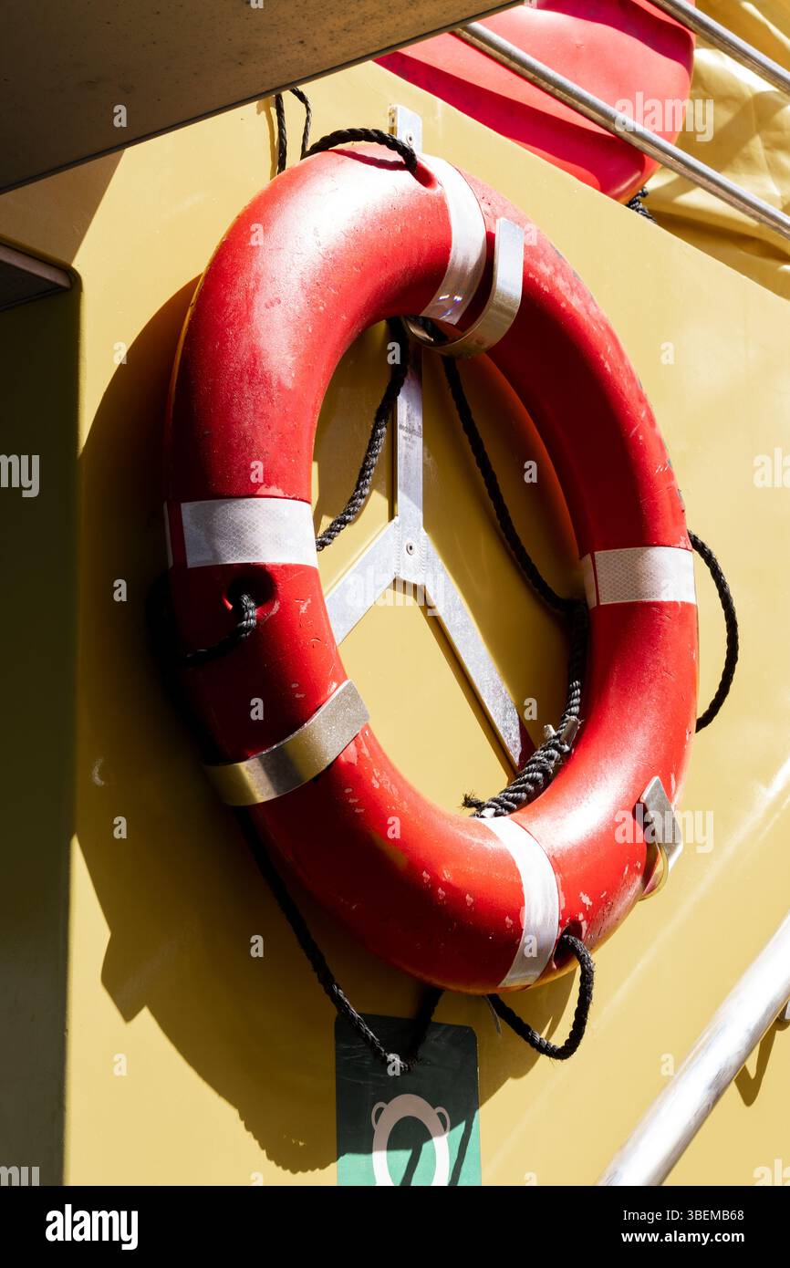 Une bouée de sauvetage rouge vif équipée de cordes, attachée à un mur jaune, contrastant fortement sous la lumière du soleil. Banque D'Images