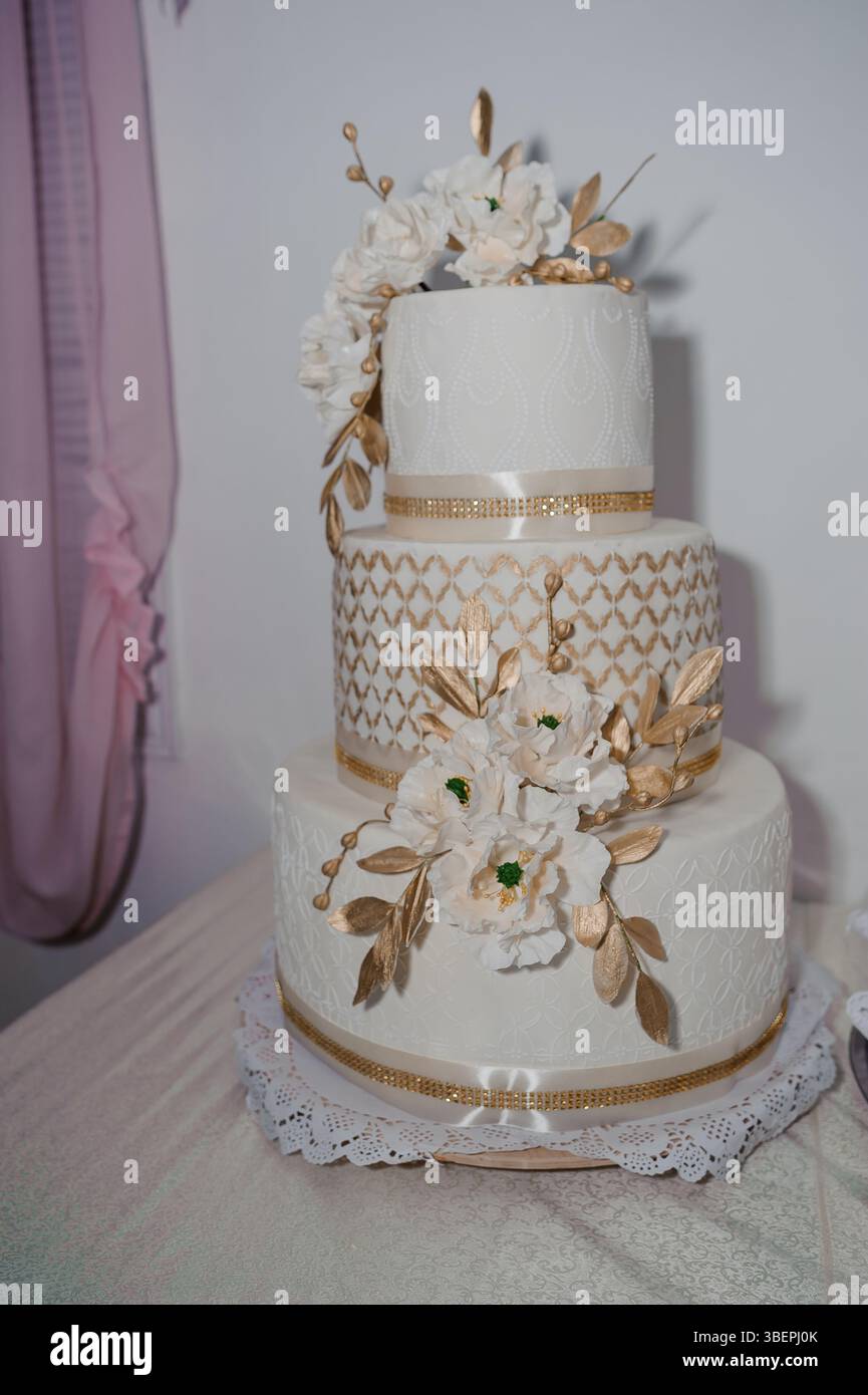 Gâteau de mariage élégant à trois niveaux avec des détails complexes de dentelle blanche, des accents dorés et des décorations florales sur un fond rose doux, célébrant un Banque D'Images