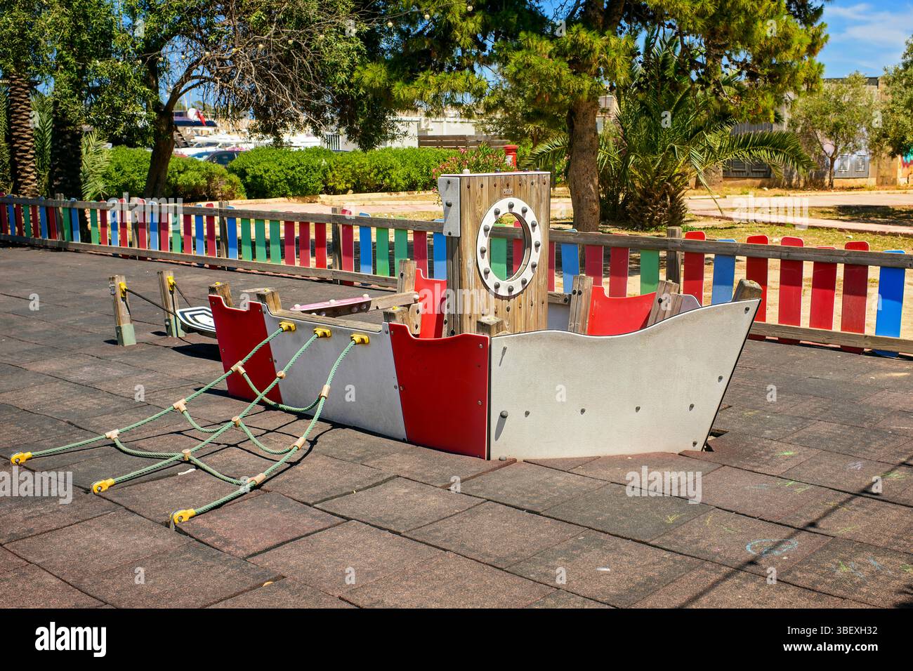Structure de jeu de style bateau pirate basse avec filets de corde dans un parc ensoleillé. Entouré par une clôture en bois colorée, des arbres et des palmiers, Banque D'Images