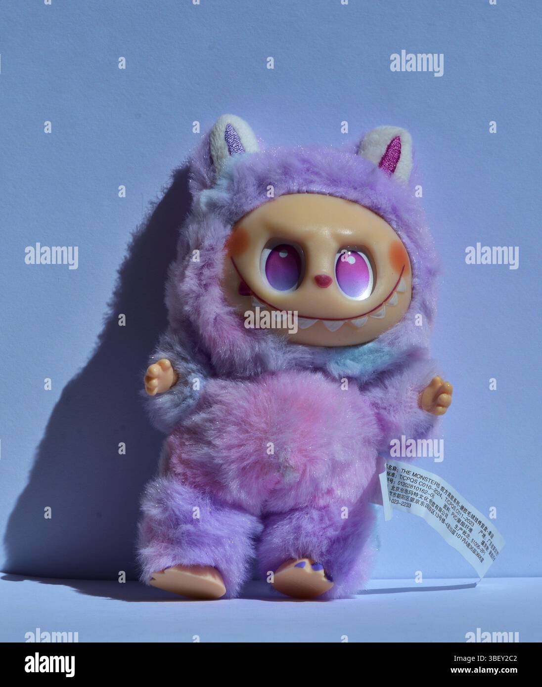 Figurine jouet Labubu ludique dans un costume de monstre violet rose moelleux avec de grands yeux roses et un sourire espiègle, posé sur un fond bleu clair Banque D'Images