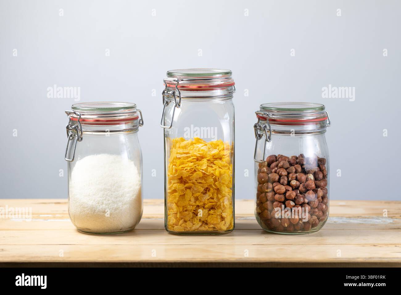 Bocaux en verre avec noix de coco desséchée, flocons de maïs et noisettes sur une surface en bois, organisés pour le stockage de la cuisine ou la préparation des aliments Banque D'Images