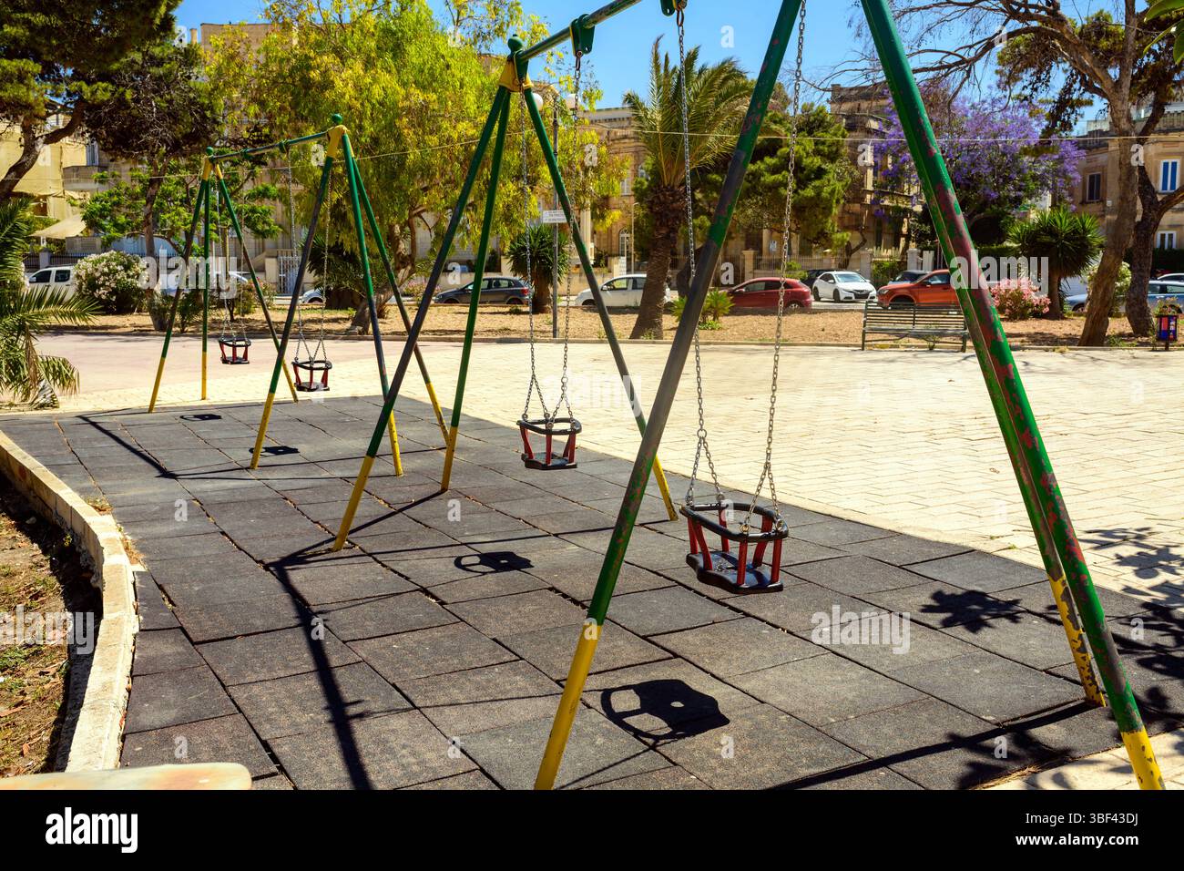 Une rangée de balançoires vides sur une aire de jeux tranquille dans un parc urbain par une journée ensoleillée. Les ombres tombent sur le tapis en caoutchouc, avec des arbres, des voitures et des bâtiments Banque D'Images