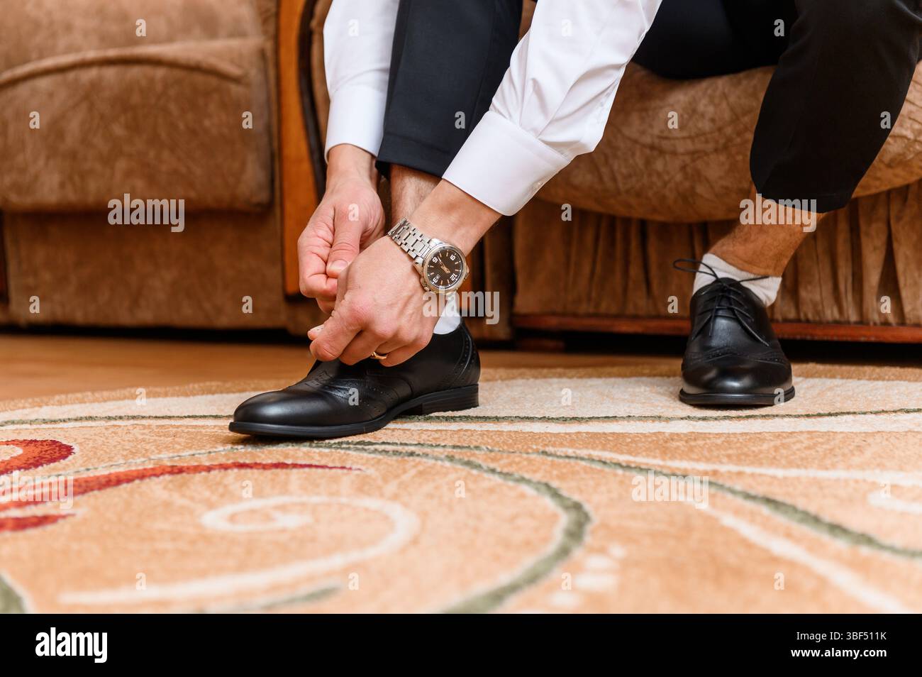 Un marié attache soigneusement ses chaussures à lacets en cuir noir tout en ajustant sa montre-bracelet, mettant en vedette une scène sophistiquée de préparation de jour de mariage avec un Banque D'Images