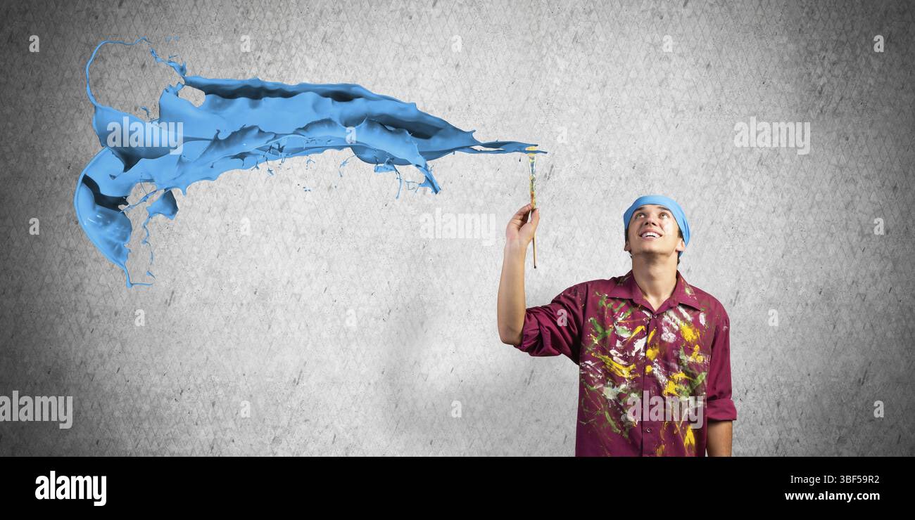 Jeune artiste peignant coup de pinceau bleu. Peintre masculin en chemise sale et bandana avec pinceau sur fond de mur gris. Passe-temps créatif et artistique Banque D'Images