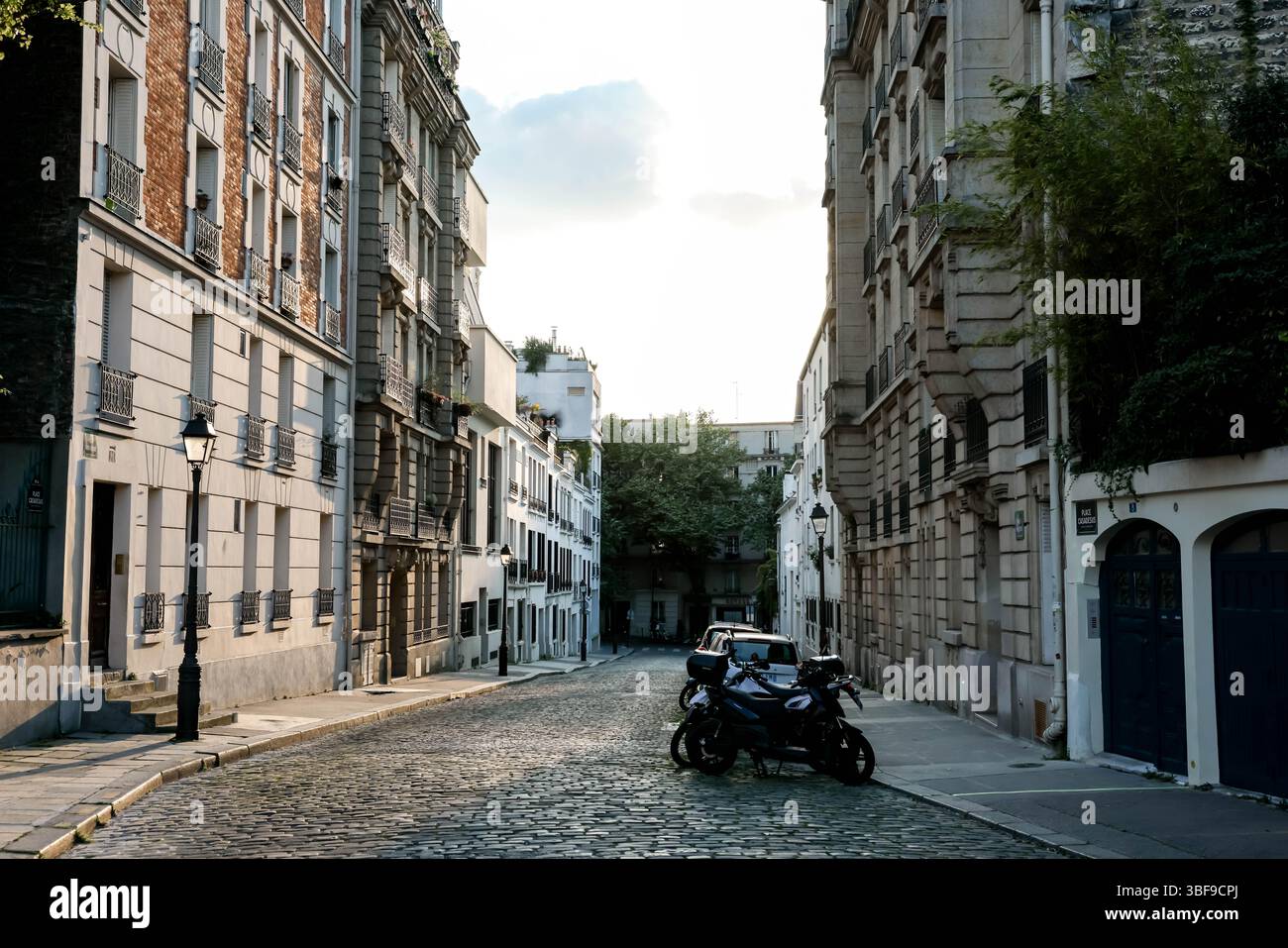 Une vue sur une rue parisienne courbe avec une surface pavée, bordée de bâtiments traditionnels, avec deux motos garées à la lumière du soir. Banque D'Images