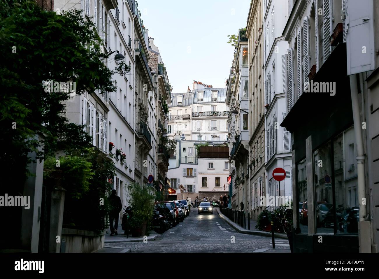 Une vue sur une rue parisienne étroite et atmosphérique bordée d'immeubles d'habitation traditionnels, de scooters garés et de phares d'une seule voiture Banque D'Images