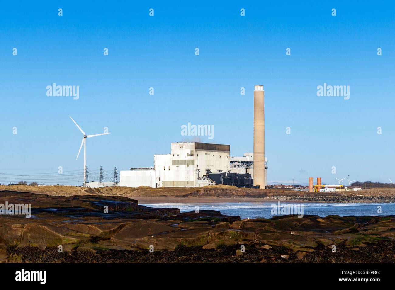 Centrale électrique côtière avec une éolienne et Smokestack par temps clair, les vagues s'écrasant sur le rivage rocheux Banque D'Images