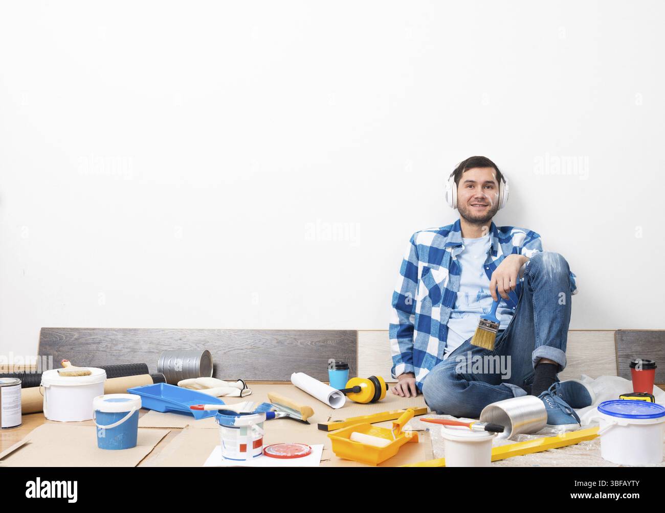 Bel homme barbu écoutant de la musique dans des écouteurs à la maison. Rénovation de la maison après le concept de déménagement. Outils de peinture et matériel sur le sol. Jeune cauca Banque D'Images