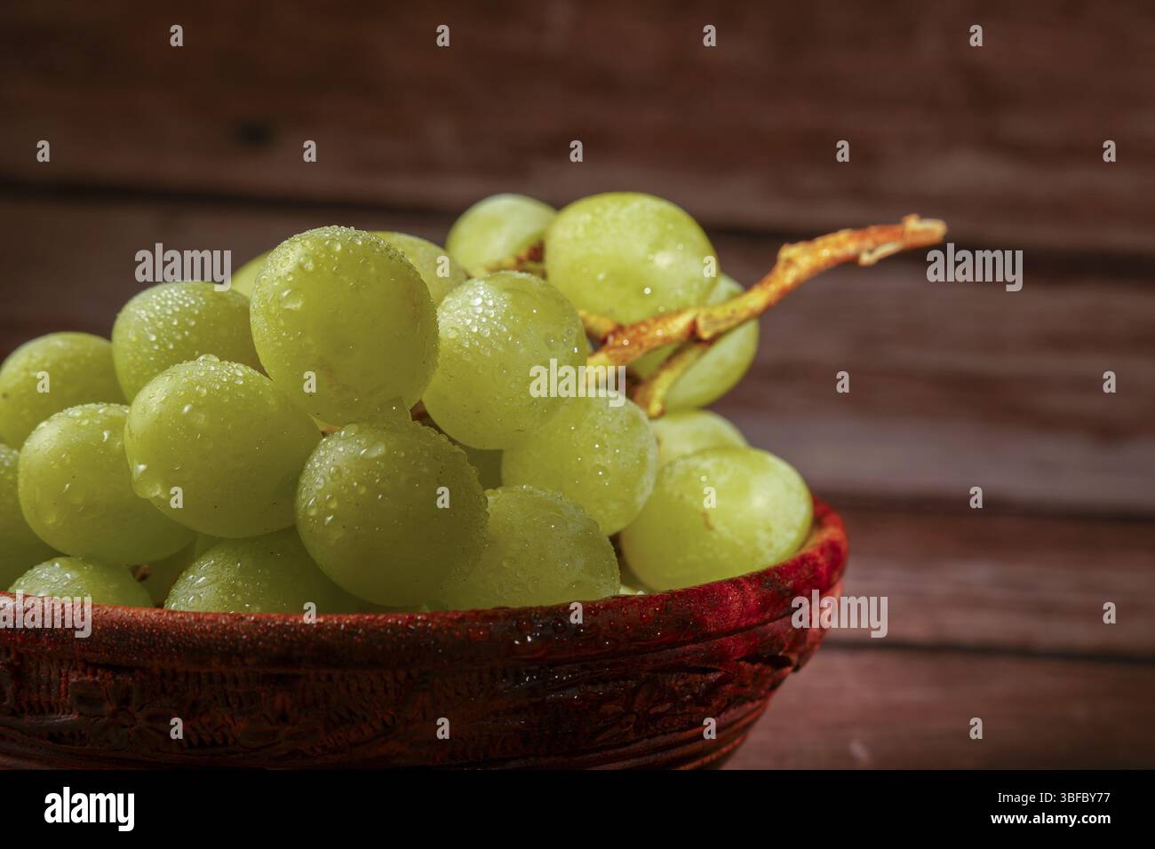 Grappe de raisins verts mûrs avec des gouttes d'eau, servis dans un bol en bois sculpté sur une table en bois Banque D'Images