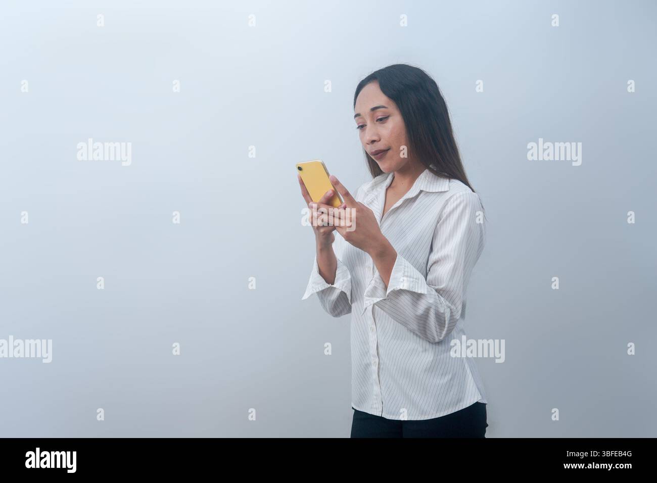 Femme indonésienne utilisant un smartphone tout en se tenant debout sur un fond blanc. Idéal pour des thèmes comme la technologie, la communication et les médias sociaux. Banque D'Images