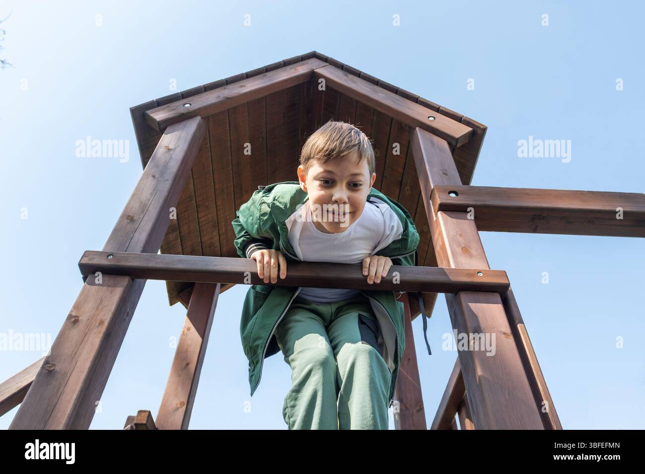 Un garçon grimpe sur une structure de jeu en bois, regardant vers le bas avec un sourire. pour des thèmes tels que les activités pour les jeunes, le temps de jeu pour les enfants ou le plaisir estival en plein air. Banque D'Images