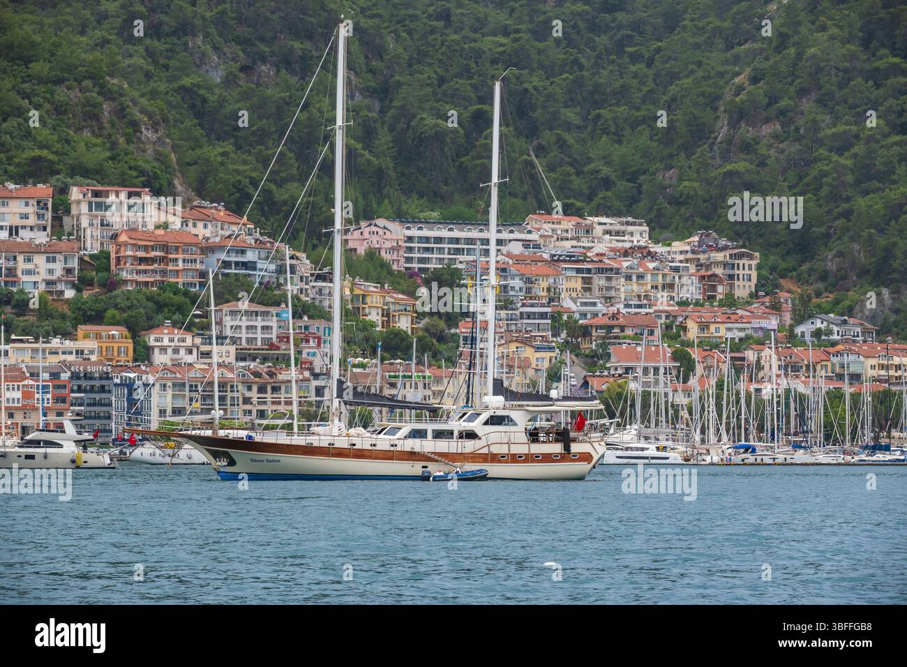 De nombreux bateaux, dont un yacht marron et blanc de premier plan, reposent dans les eaux portuaires de Fethiye, Türkiye, sur fond de bâtiments à flanc de colline. Banque D'Images
