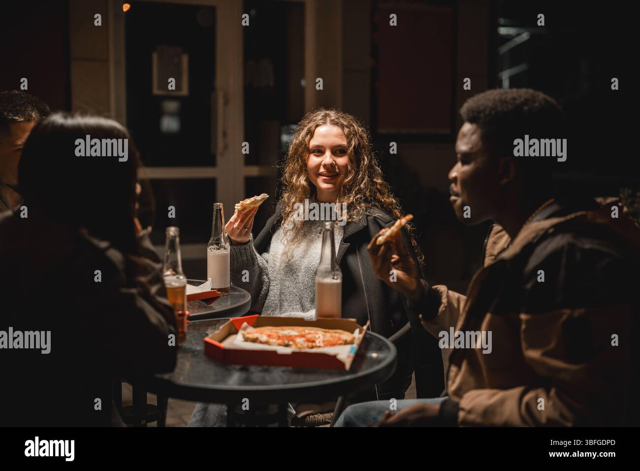Groupe d'amis appréciant pizza et boissons la nuit dehors dans un cadre urbain confortable Banque D'Images