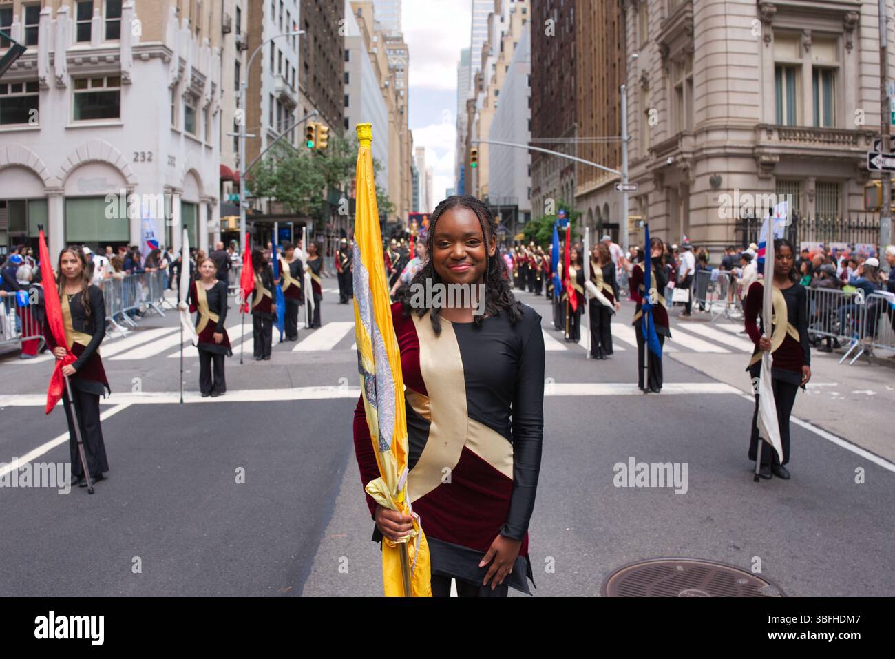 Une jeune femme dans un uniforme de garde de couleur noir et Bordeaux sourit, tenant un drapeau jaune et or. D'autres membres de garde de couleur, dans des uniformes similaires, le sont Banque D'Images