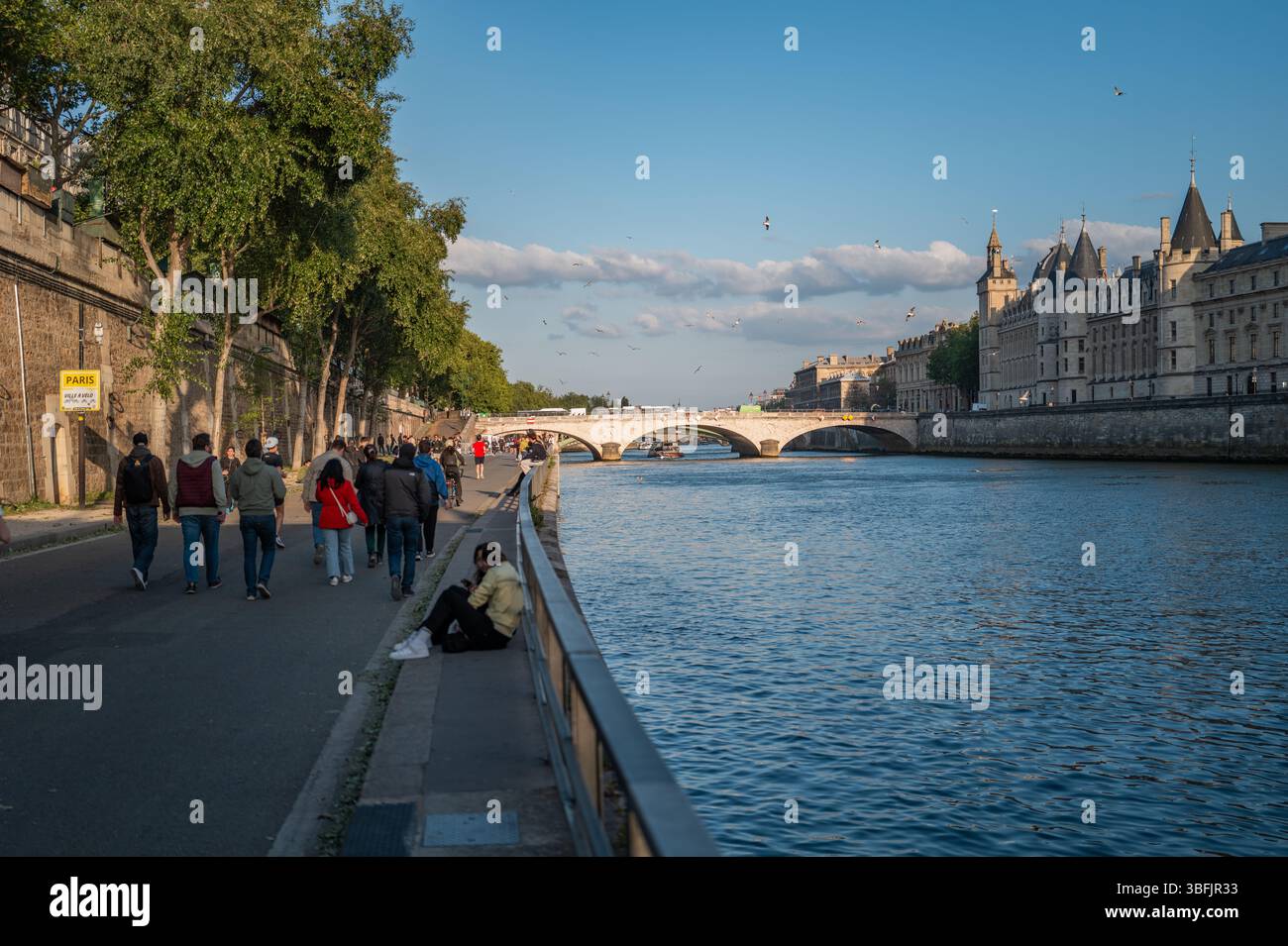 Les gens marchant et assis le long de la promenade de la Seine à Paris pendant un après-midi ensoleillé avec des bâtiments historiques en arrière-plan. Banque D'Images