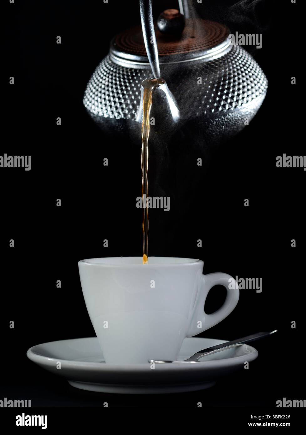 Boisson chaude versée à partir d'une théière argentée dans une tasse blanche sur fond noir, créant une atmosphère chaleureuse et accueillante Banque D'Images