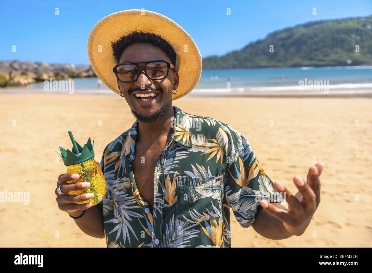 Jeune homme avec chapeau de paille et chemise hawaïenne souriant et tenant un cocktail d'ananas sur une plage ensoleillée Banque D'Images