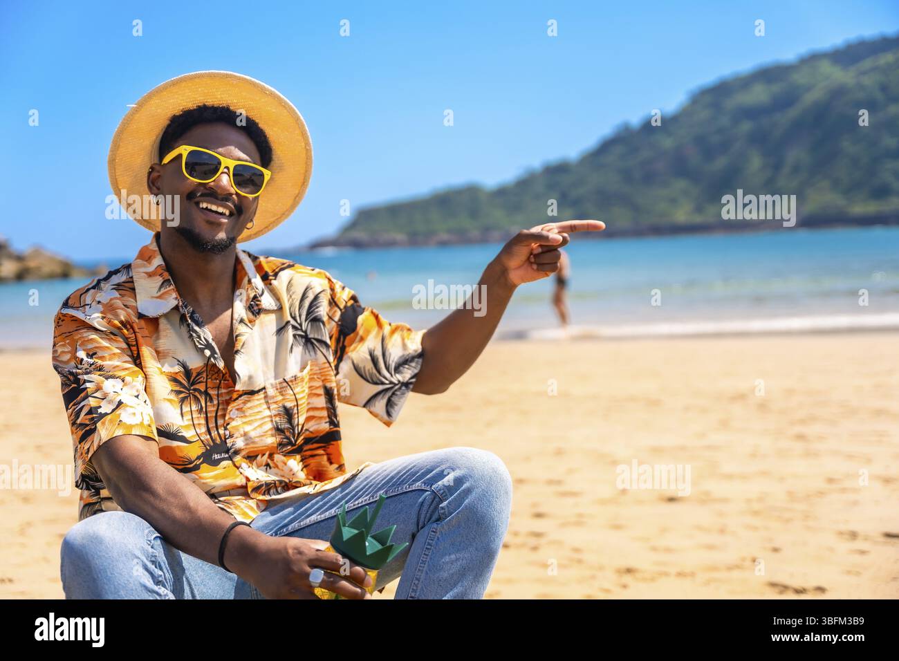 Homme noir portant une chemise hawaïenne, des lunettes de soleil et un chapeau de paille pointant son doigt sur quelque chose sur une plage tropicale Banque D'Images