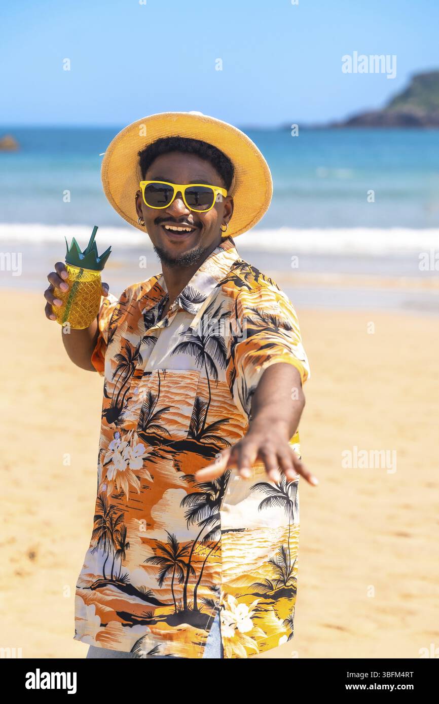 Jeune homme portant une chemise hawaïenne et des lunettes de soleil dégustant un cocktail d'ananas sur une plage ensoleillée Banque D'Images
