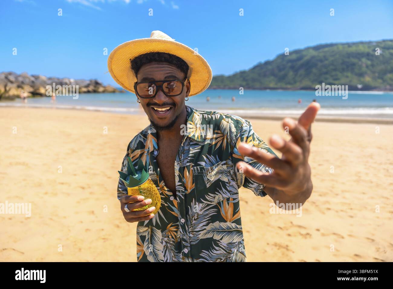 Homme noir portant une chemise hawaïenne et des lunettes de soleil profitant des vacances d'été boire un cocktail sur une plage tropicale Banque D'Images
