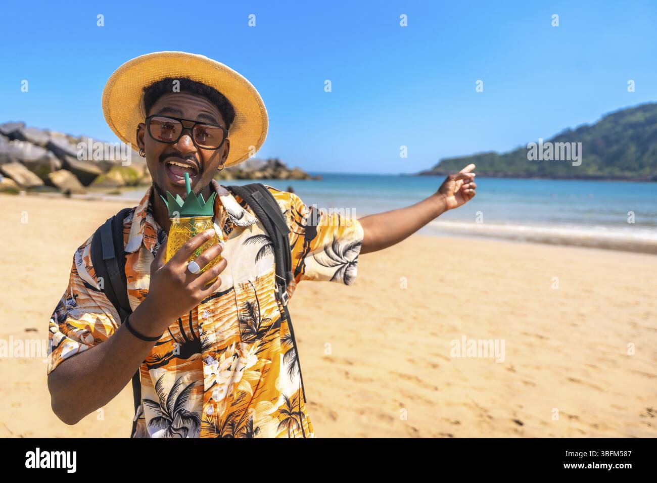 Jeune homme noir avec chapeau de paille et chemise hawaïenne buvant un cocktail sur une plage tropicale tout en pointant quelque chose Banque D'Images