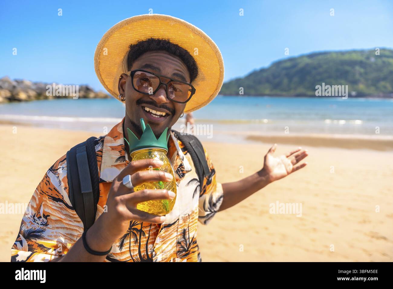 Jeune homme portant une chemise hawaïenne et un chapeau de paille buvant un cocktail d'ananas sur une plage tropicale Banque D'Images