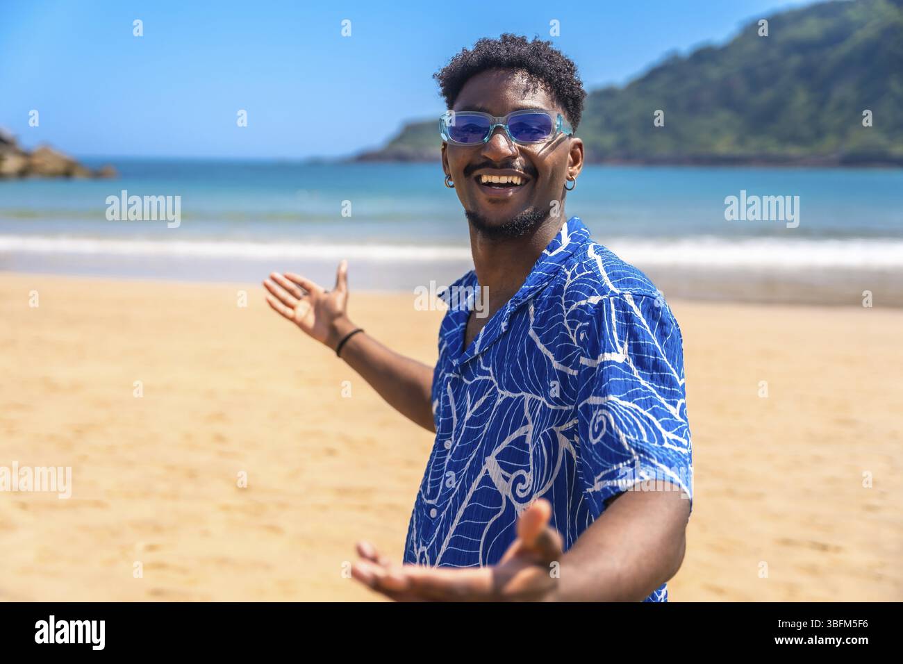 Jeune homme noir avec des lunettes de soleil et chemise hawaïenne profitant des vacances d'été accueillant les gens sur une plage tropicale à bras ouverts Banque D'Images