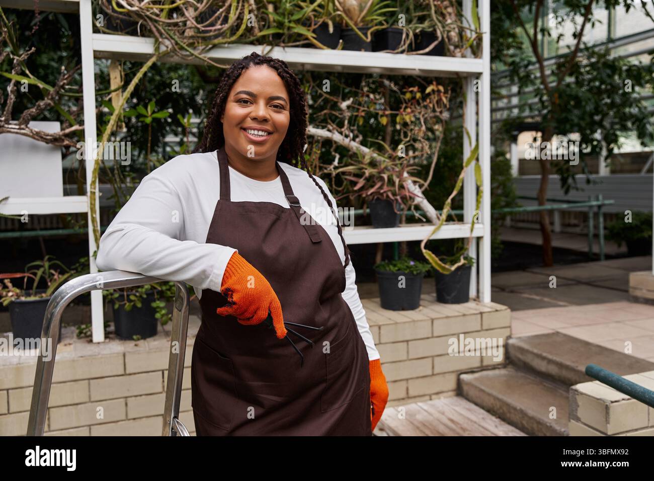 Une femme afro-américaine avec des tresses rayonne de joie tout en s'occupant des plantes dans une serre. Banque D'Images
