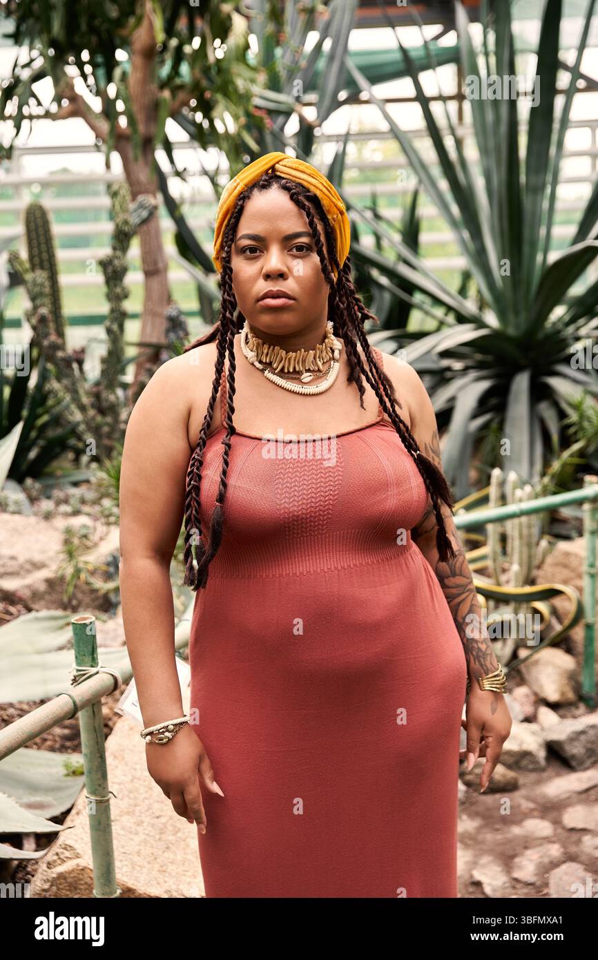 Une femme afro-américaine élégante avec des tresses se tient en toute confiance au milieu de belles plantes à l'intérieur. Banque D'Images