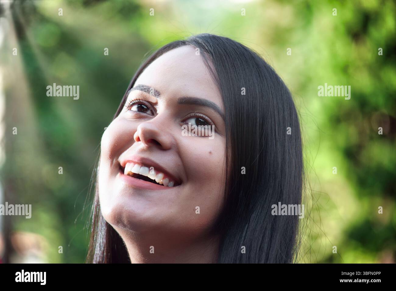 Portrait en gros plan d'une jeune femme joyeuse aux longs cheveux foncés souriant et levant les yeux. L'arrière-plan est rempli de nature verte douce, donnant l'image Banque D'Images
