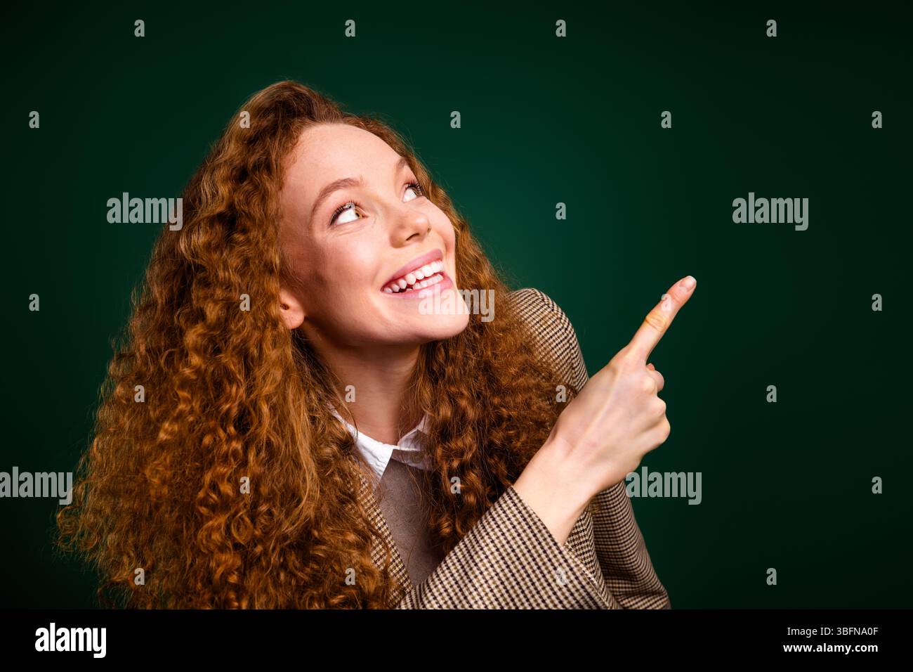 Femme d'affaires souriante aux cheveux bouclés pointant vers le haut avec sa main sur un fond vert Banque D'Images
