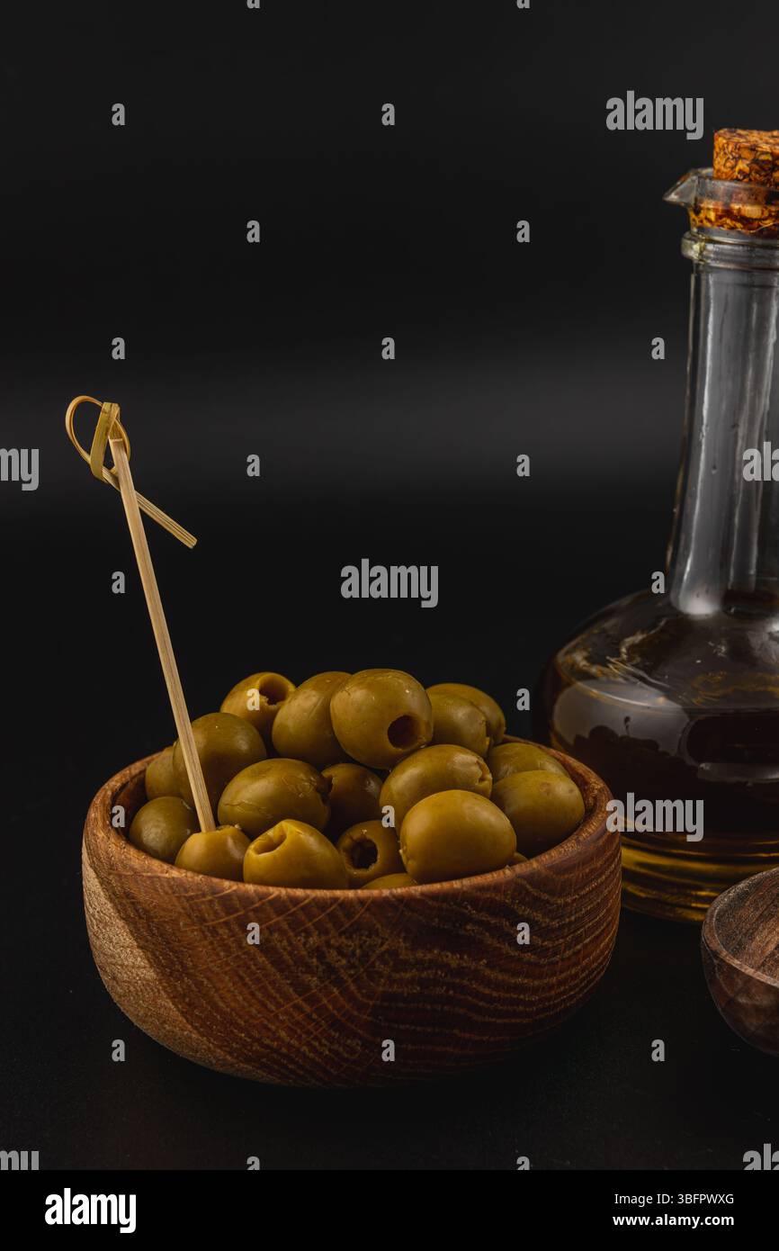 Un bol en bois rempli d'olives vertes se trouve à côté d'une bouteille en verre d'huile d'olive. Le riche bois brun contraste avec les olives vertes vibrantes dans un dimly Banque D'Images