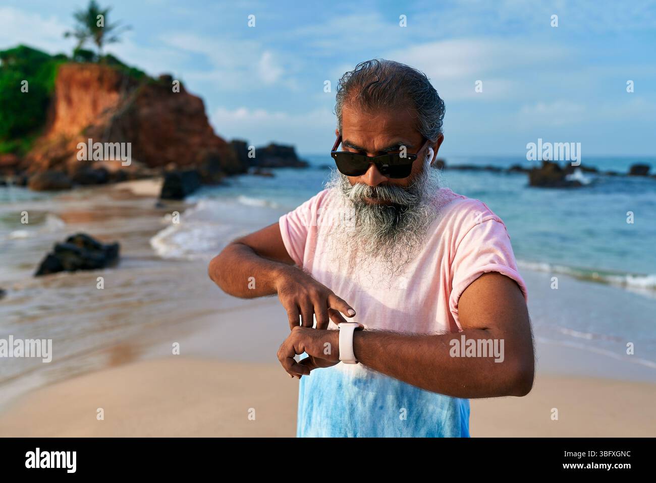 Homme ethnique senior avec la barbe grise sur la plage utilise une montre intelligente et des écouteurs pour le fitness. Les hommes âgés aiment la technologie pour la formation, le bien-être. Le vieil homme est entré Banque D'Images