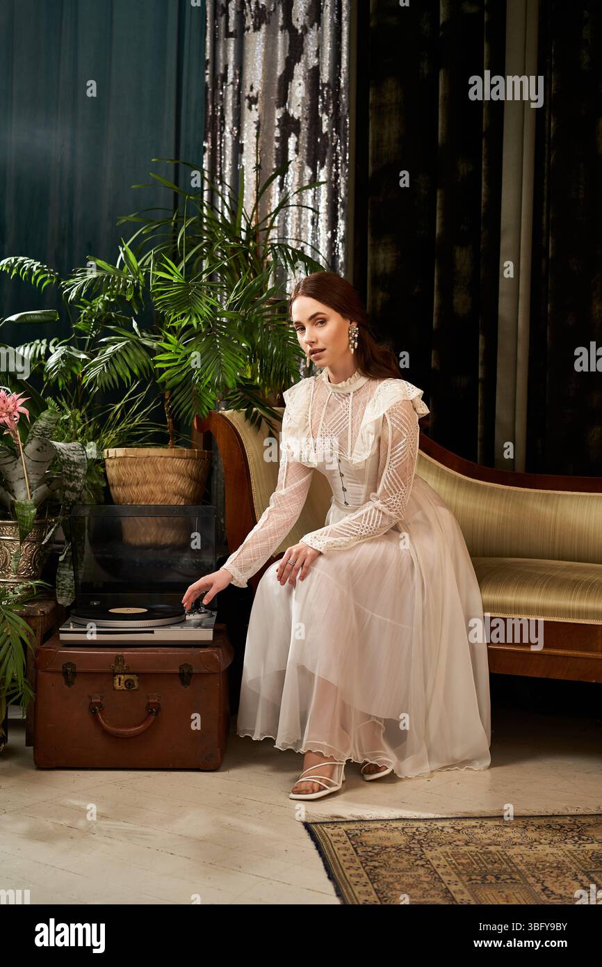 Une belle femme s'assoit gracieusement, portant une robe vintage, entourée de plantes et d'un décor classique. Banque D'Images