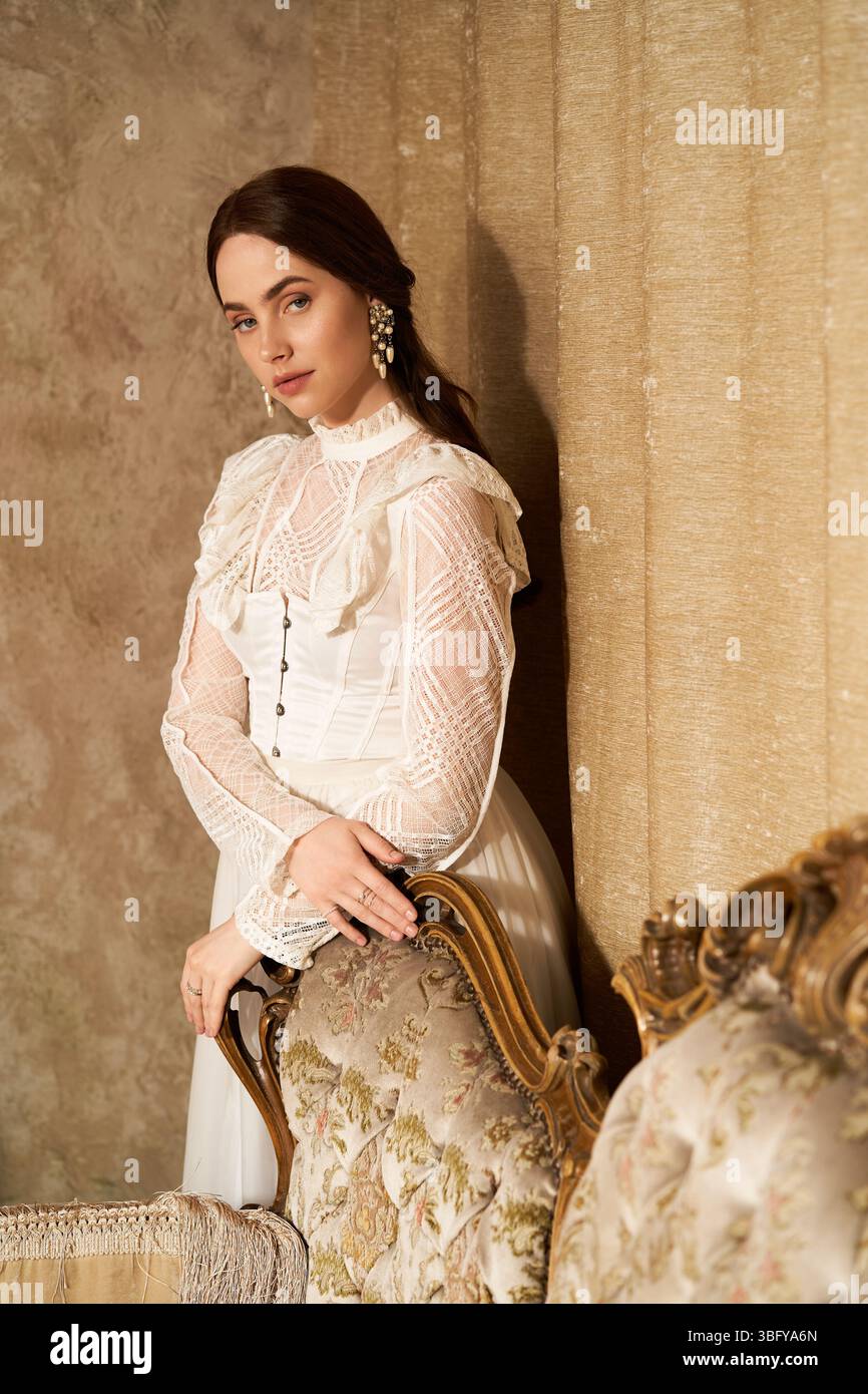 Jeune femme dans une robe vintage se penche gracieusement contre une chaise ornée, exsudant l'élégance. Banque D'Images