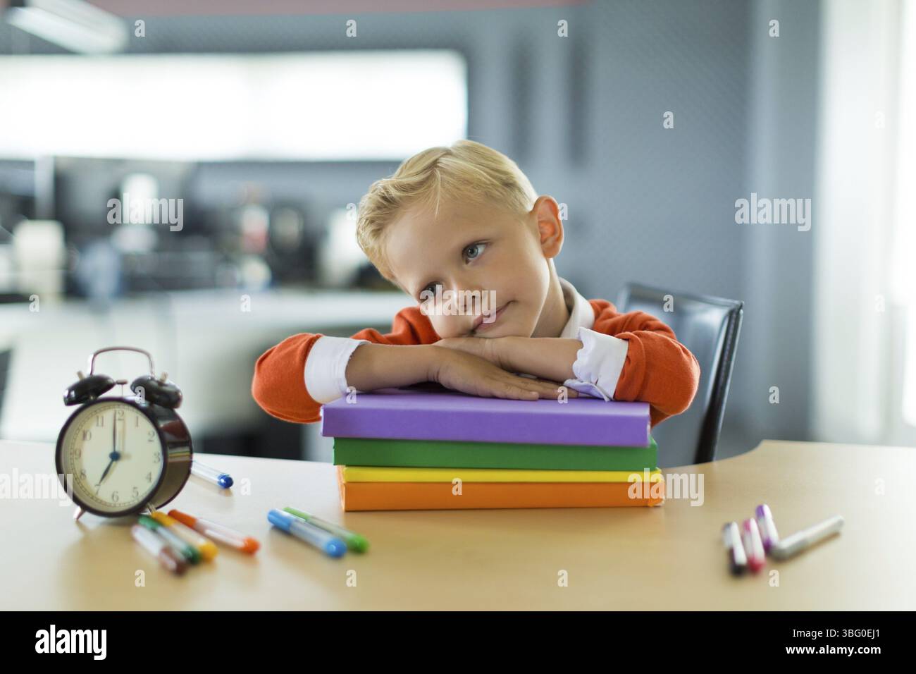 Mignon enfant caucasien blond assis à la table avec des dossiers colorés et un réveil et repose sur les dossiers colorés, fond de bureau flou ennuyé, Banque D'Images