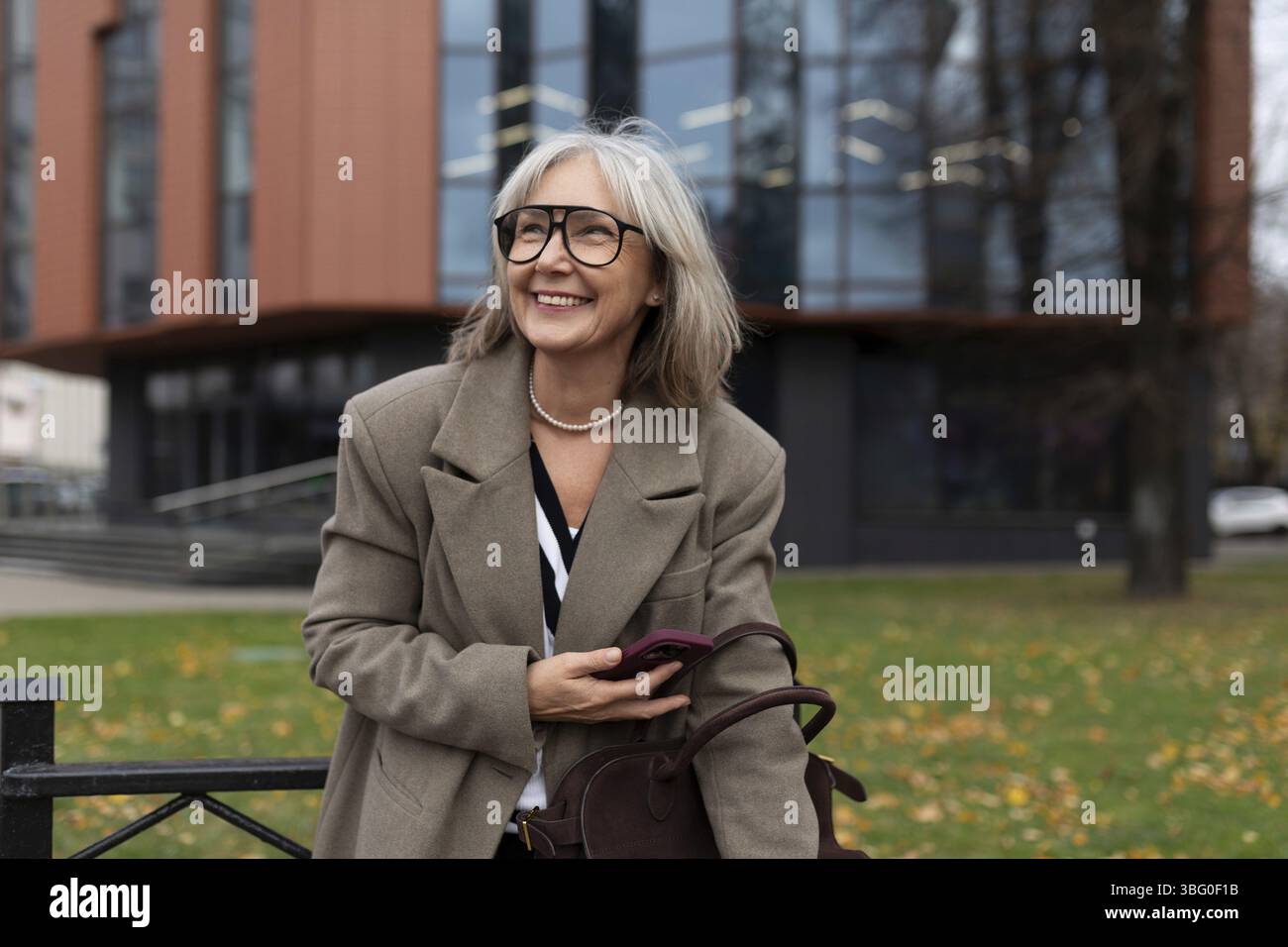 La femme d'affaires senior se tient à l'extérieur d'un immeuble de bureaux contemporain, tenant son sac et souriant chaleureusement. Elle reflète la confiance et la joie dans son pro Banque D'Images