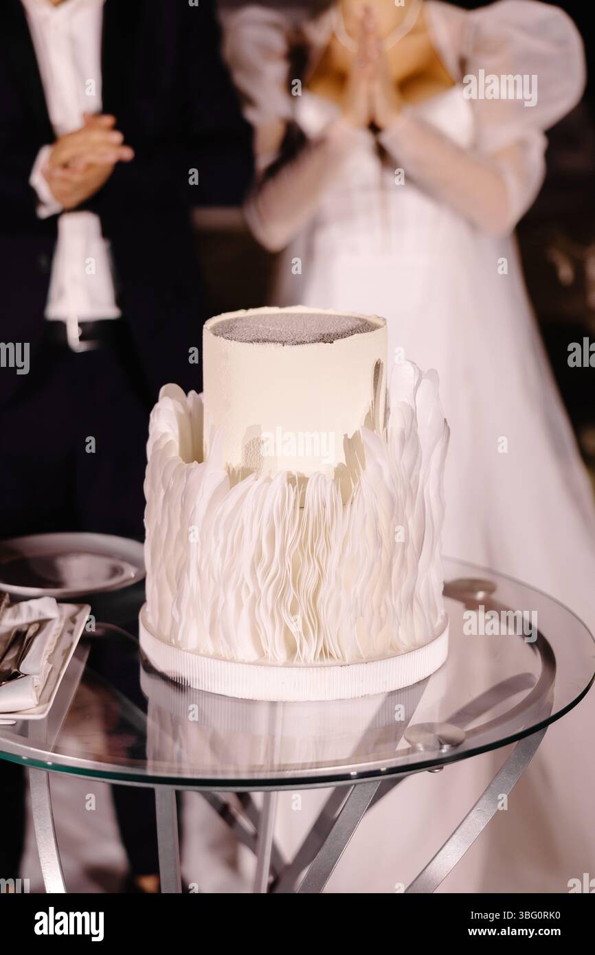 Délicieux gâteau de mariage blanc décoré avec des éléments argentés et blancs sur une table en verre, avec des applaudissements de mariée et de marié en arrière-plan Banque D'Images