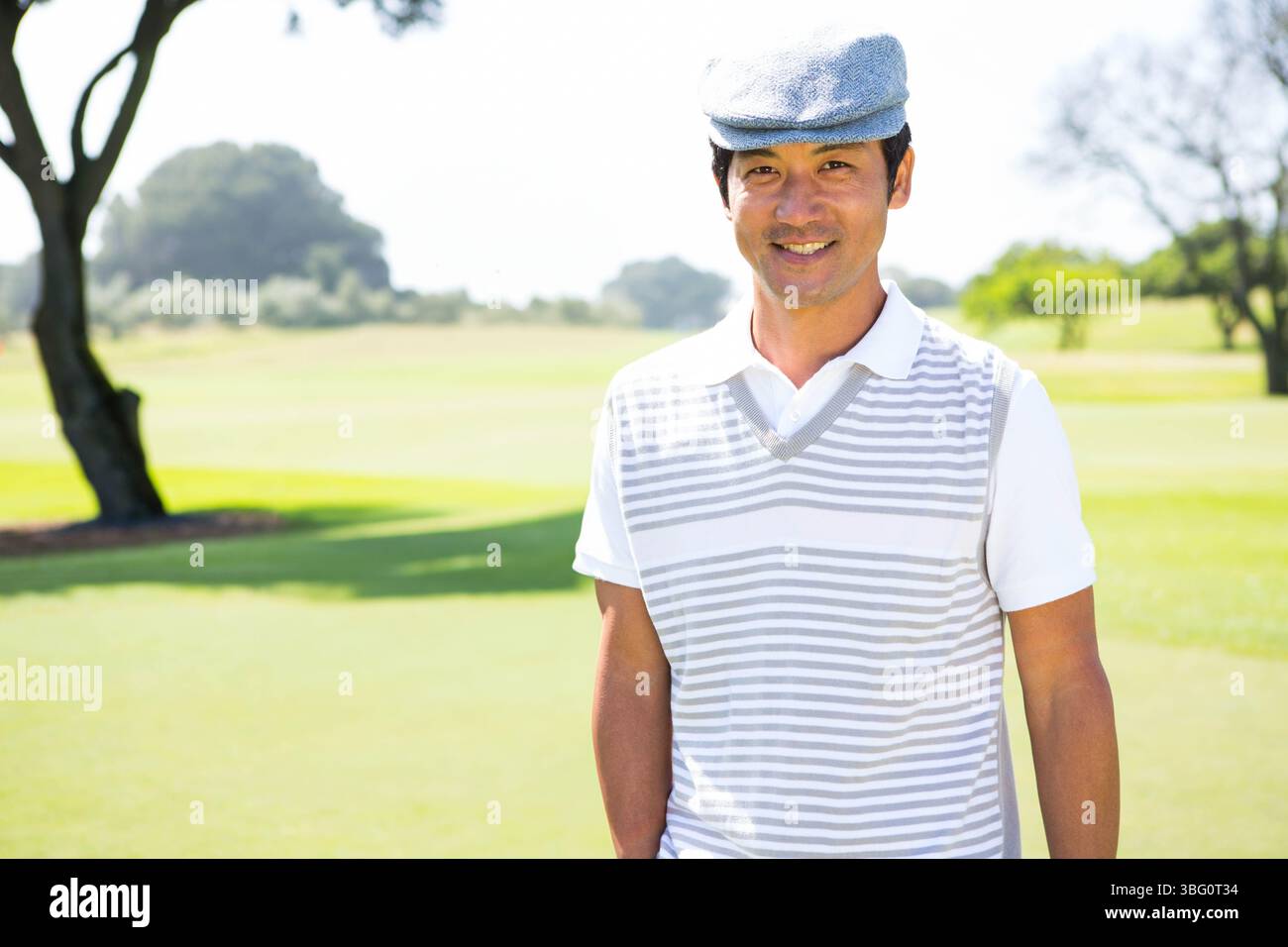 Homme japonais debout sur le fairway de golf sous la lumière du soleil portant un gilet rayé à casquette plate, espace copie Banque D'Images