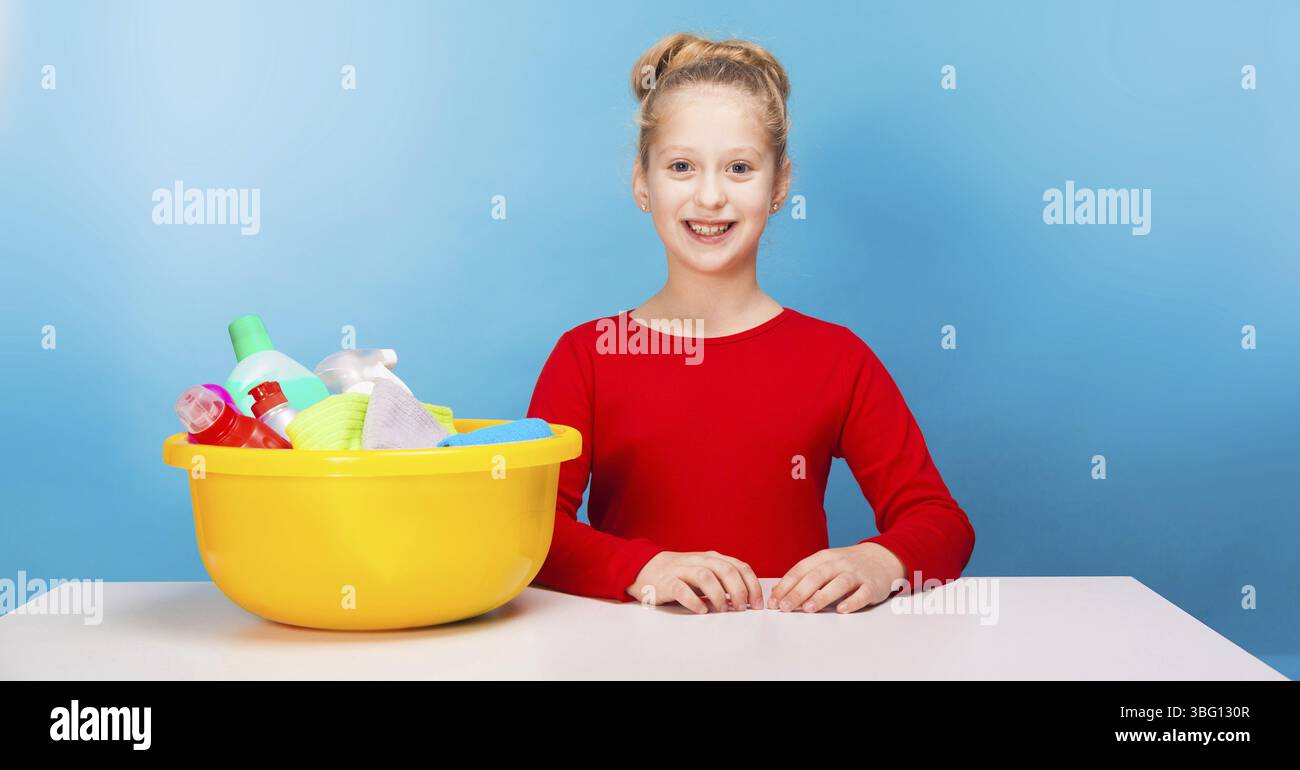 Isolé sur bleu, fille souriante en pull rouge avec updo aux cheveux clairs, assis à la table blanche avec un lavabo au citron plein de détergents multicolores Banque D'Images