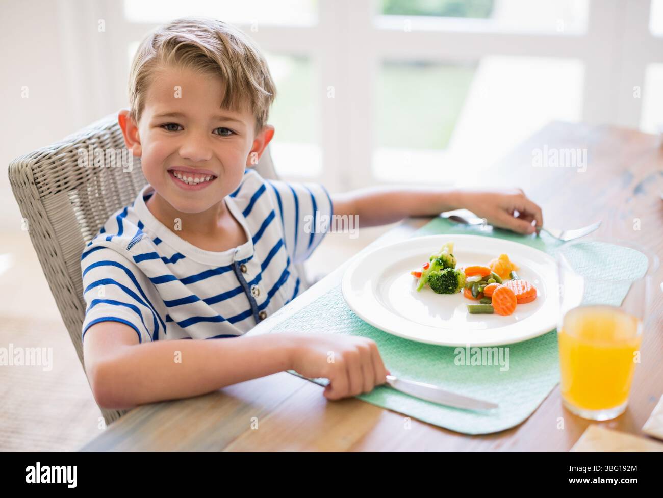 Enfant mangeant des légumes cuits à la vapeur avec une fourchette et un couteau et buvant du jus d'orange à la table à manger à la maison Banque D'Images