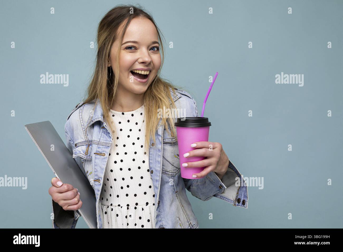 Image d'une joyeuse femme blonde européenne souriante et debout contre un mur bleu isolé avec un carnet argenté et un café à emporter dans les mains Banque D'Images