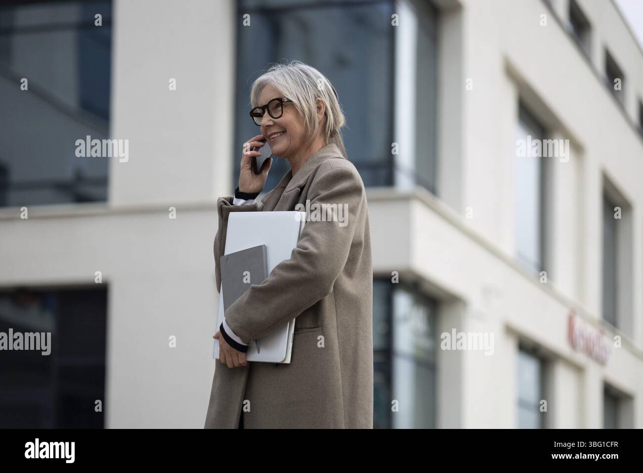 Une femme d'affaires senior s'est engagée dans une conversation téléphonique, portant un ordinateur portable et vêtue d'un manteau professionnel. Elle semble confiante en dehors d'un mépris Banque D'Images
