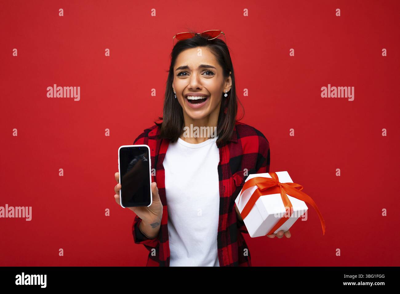 Schoene positive staunende Junge brunet Frau isoliert ueber roten Hintergrund Wand mit weissen laessigen T-shirt und rote und schwarze shirt mit weiss Banque D'Images