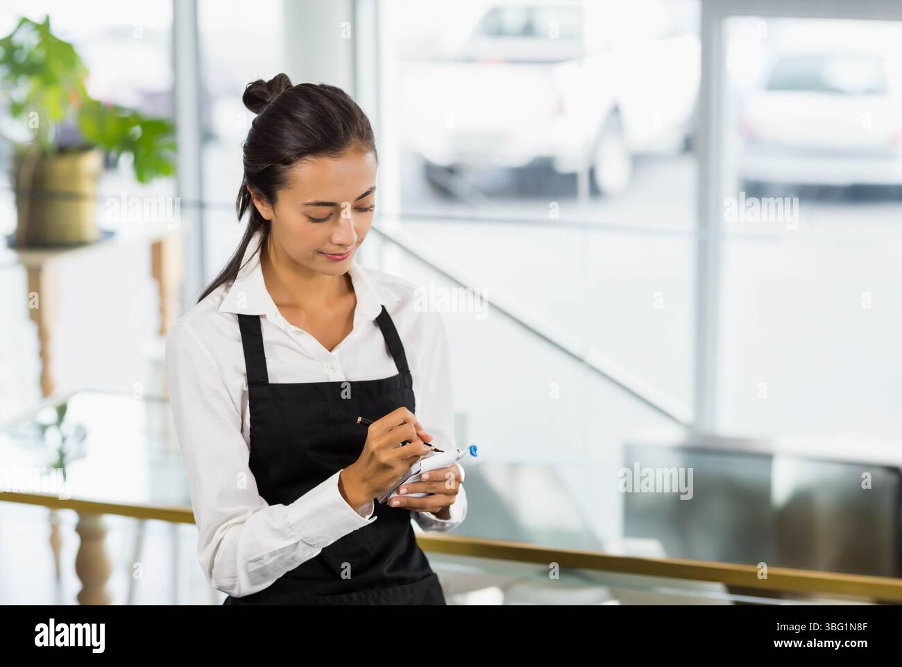 Serveuse asiatique debout à la rampe du restaurant portant un tablier noir ordre d'écriture avec un stylo sur le bloc-notes Banque D'Images