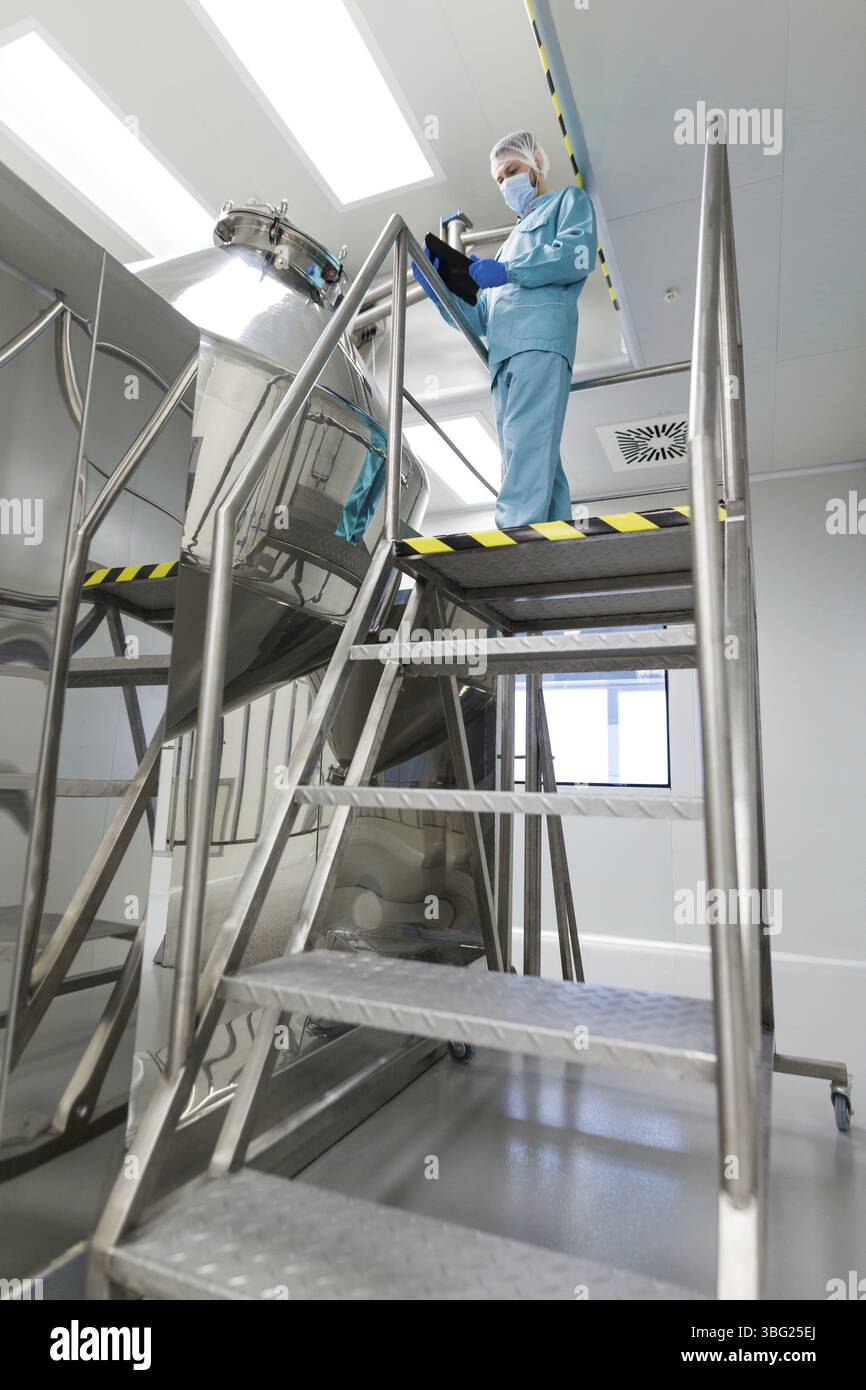 Image verticale, scientifique caucasien en costume de laboratoire bleu et gants se tiennent sur une échelle en acier et prennent des notes sur le réservoir chromé Banque D'Images