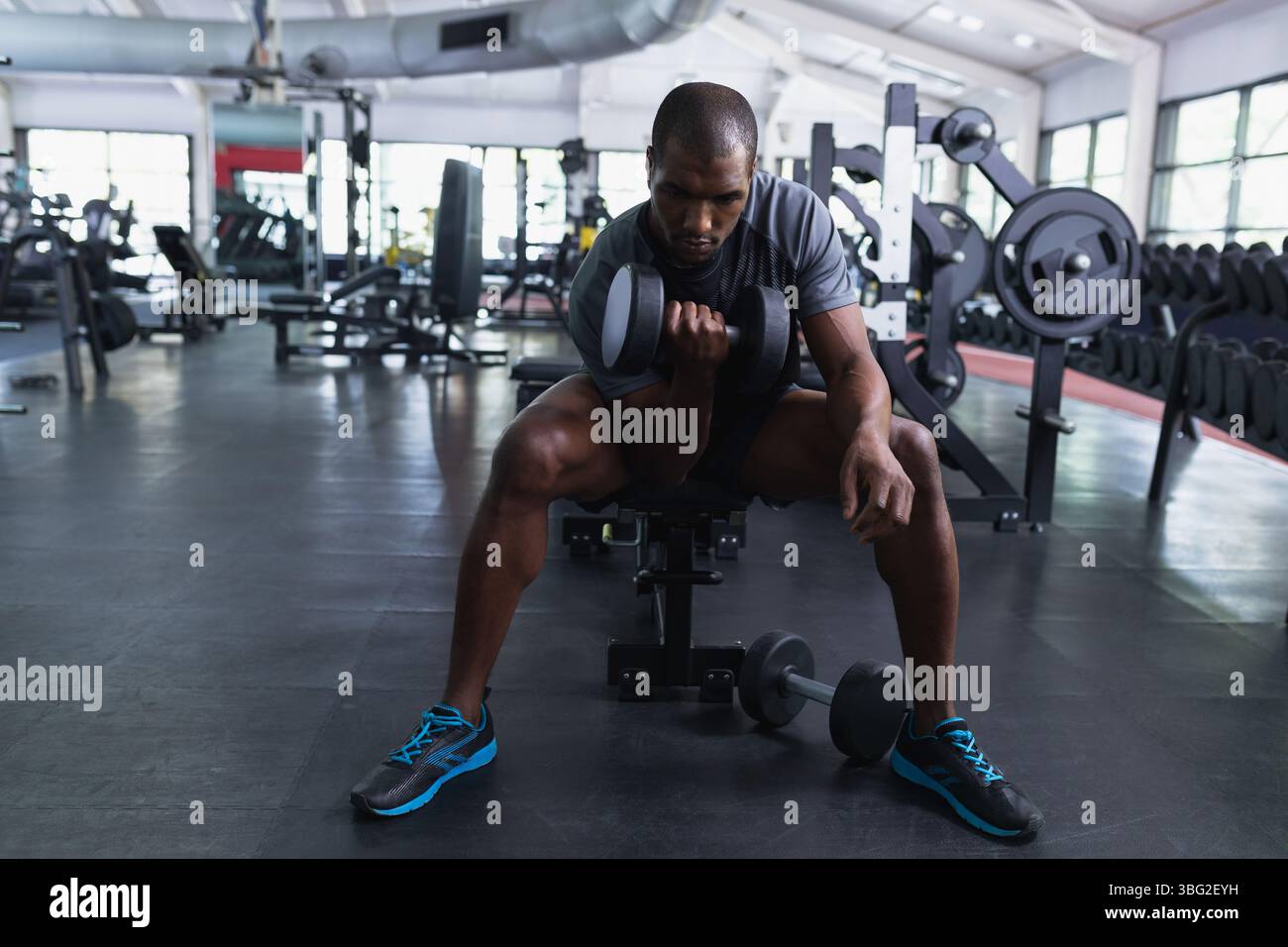 Homme assis sur le banc curling haltères à l'intérieur du gymnase avec station de presse de banc et rangées d'haltères Banque D'Images