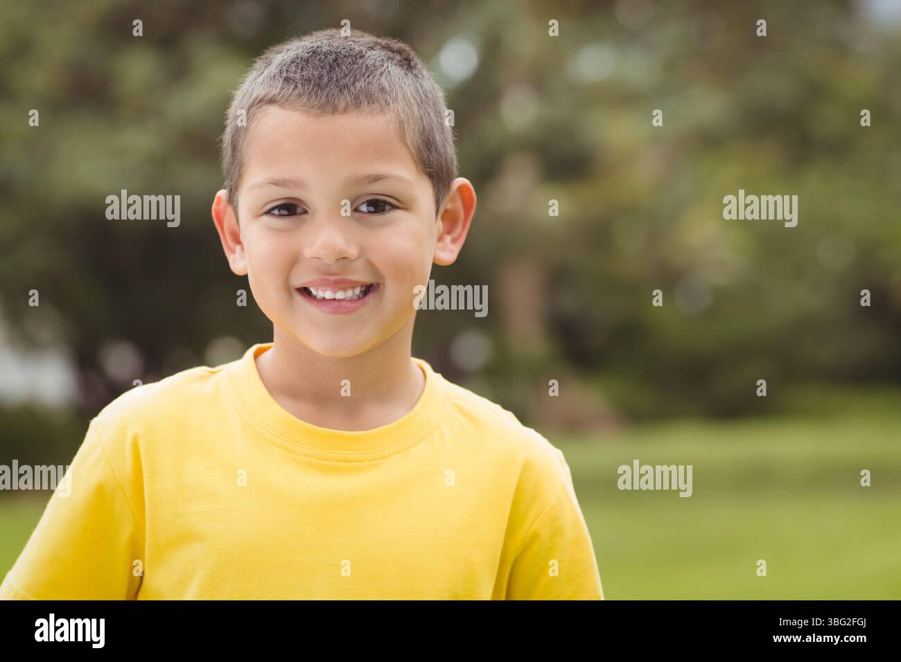 Garçon souriant portant un T-shirt jaune debout dans le parc ensoleillé avec l'herbe floue et le feuillage Banque D'Images