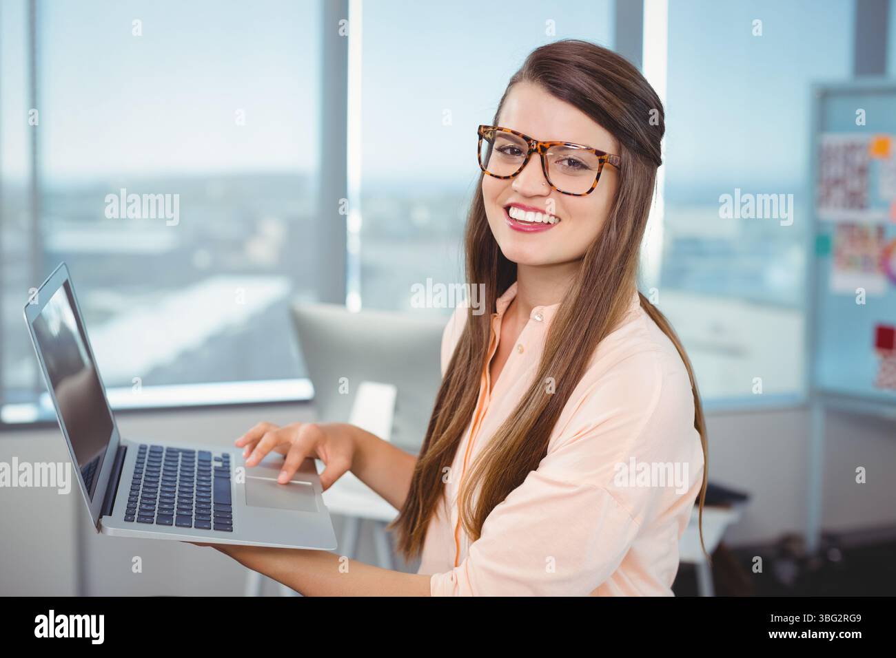 Travailleuse de bureau tenant un ordinateur portable argenté près de la fenêtre portant des lunettes par tableau d'affichage, espace de copie Banque D'Images