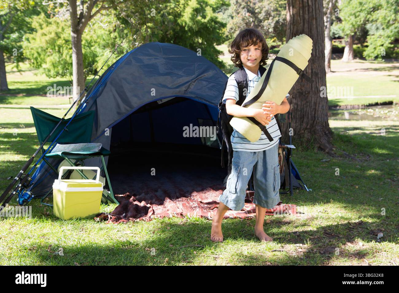 Garçon portant un coussin de couchage roulé et un sac à dos devant la tente bleue au parc, espace de copie Banque D'Images